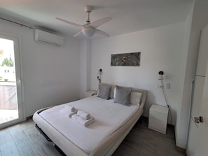 A bedroom with a bed, a ceiling fan, an air conditioning unit, a bedside table, a nightstand, and a painting on the wall.