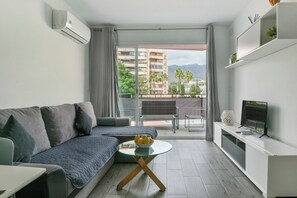 A modern living room with a sofa, a glass coffee table, a TV, and a balcony with a view of buildings and palm trees.