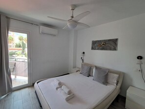 A hotel room with a bed, a ceiling fan, an air conditioning unit, a painting on the wall, and a view of the outside through a sliding door.