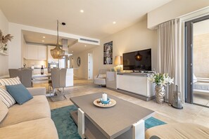 A modern living room with a sofa, coffee table, and a TV. There is a kitchen in the background with cabinets and a dining area.