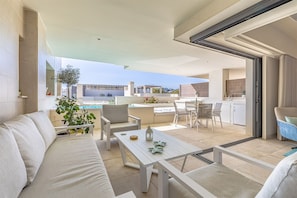 A modern outdoor living area with a sofa, chairs, a table, and a view of the pool and kitchen.