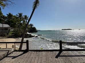 A wooden pier with a railing, overlooking a beach with palm trees and a view of the ocean.