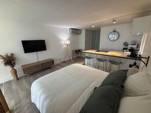 A modern bedroom with a large bed, a TV mounted on the wall, a small kitchen area with a coffee machine, and a bedside table.