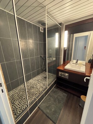 A modern bathroom with a glass-enclosed shower, dark tiles, a wooden vanity, and a large mirror.