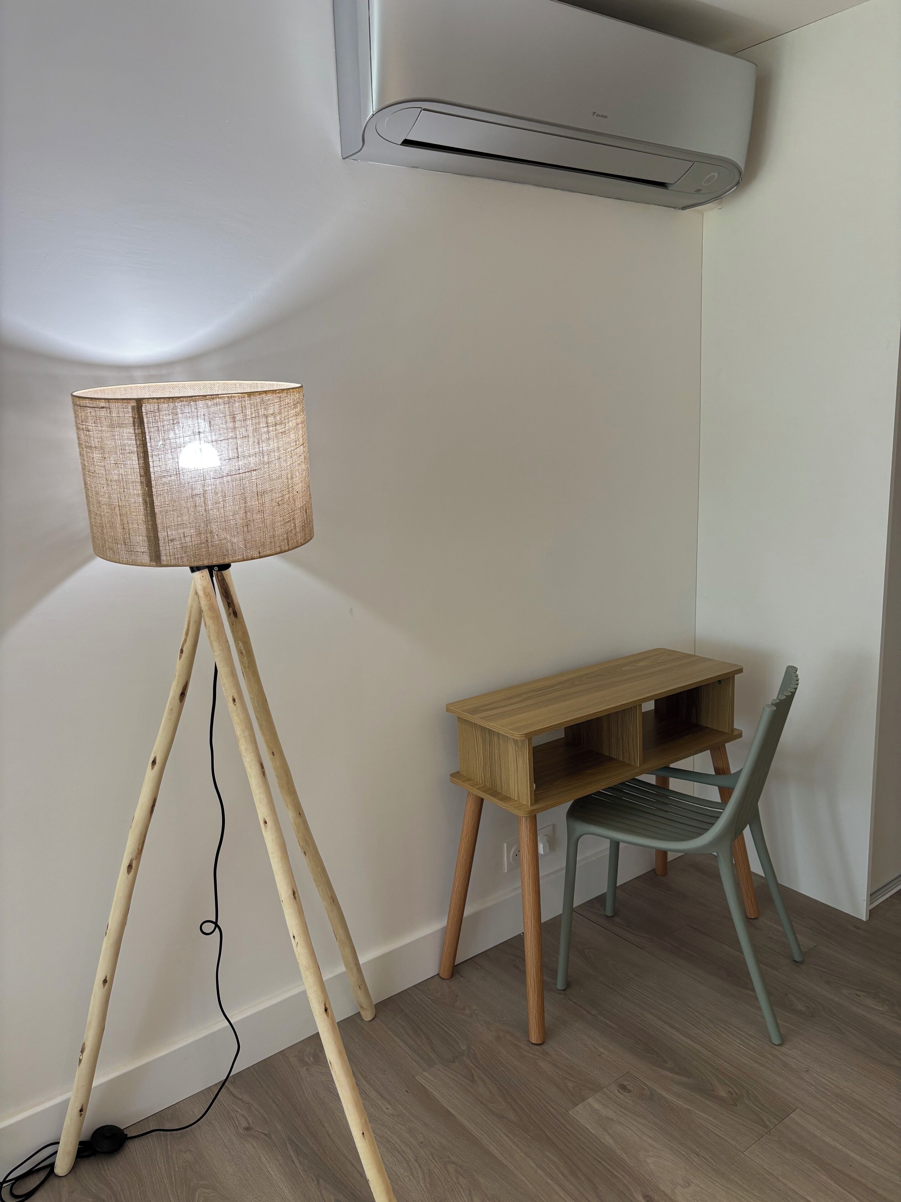 A corner room with a wooden floor, a small wooden desk, a green chair, and a floor lamp.
