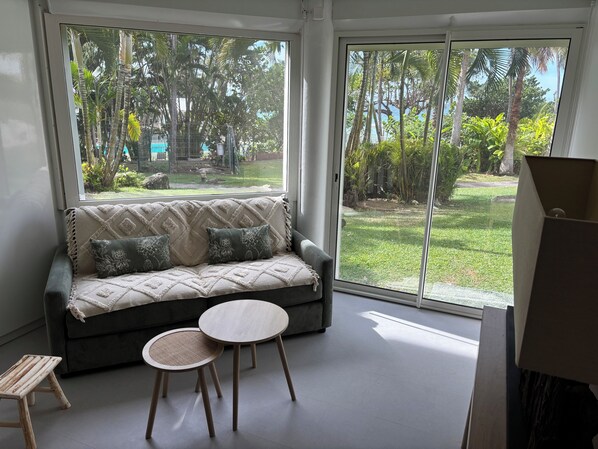A modern living room with a sofa, two round tables, and a view of a lush garden through large windows.