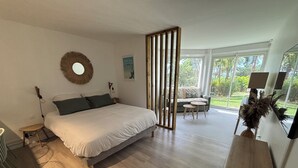 A modern bedroom with a large bed, a small table, a sofa, and a view of a garden through large windows. - Spacious studio on the seafront, newly renovated (Le Gosier)