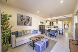 Een moderne woonkamer met een bank, salontafel en voetenbankje, een eethoek met stoelen, en een keuken op de achtergrond.