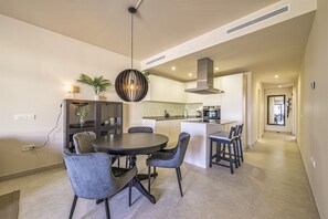 A modern kitchen with a central island, dining area with round table and chairs, and a hallway leading to other rooms.