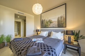 A bedroom with a large bed, a bedside table with a lamp, a wall-mounted artwork, and a potted plant.