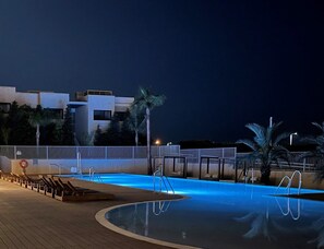 En swimmingpool med træterrasse, palmer og en moderne bygning i baggrunden.