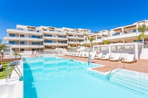 Ein Poolbereich eines Hotels mit mehreren Balkonebenen, Liegestühlen und einem wolkenlosen, blauen Himmel.