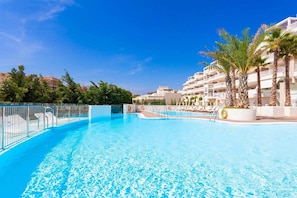 A clear blue swimming pool with lounge chairs and palm trees, surrounded by multi-story buildings.