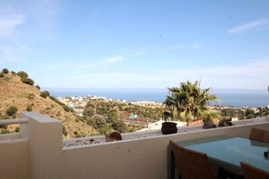 A terrace with a glass table and chairs, overlooking a coastal landscape with hills and a view of the sea.