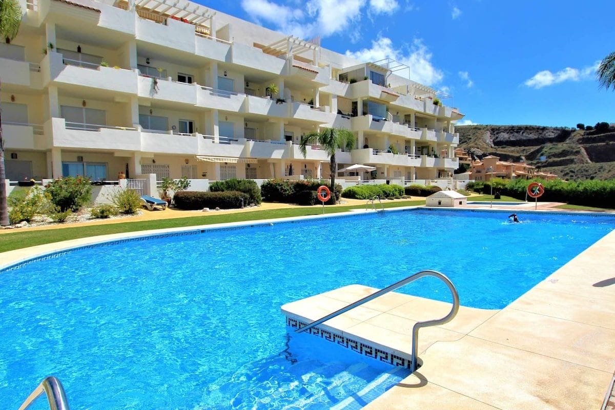 A swimming pool with a clear blue water, surrounded by a building with multiple balconies and a hilly landscape in the background.
