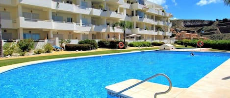 A swimming pool with a clear blue water, surrounded by a building with multiple balconies and a hilly landscape in the background.