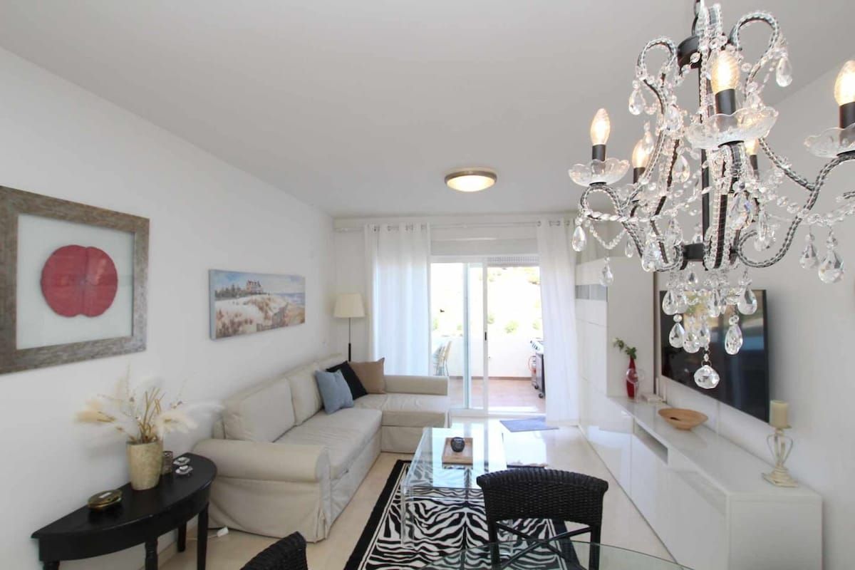 A modern living room with a chandelier, a sofa, a glass coffee table, and a television.