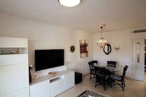 A living room with a flat-screen TV, a dining table with chairs, a chandelier, and a white cabinet.