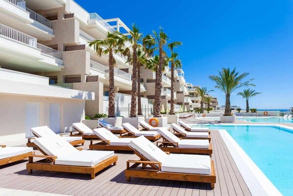 A poolside area with sun loungers, palm trees, and a clear blue sky.
