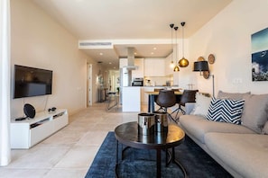 A modern living room with a sofa, a coffee table, and a TV. There is a kitchen in the background.