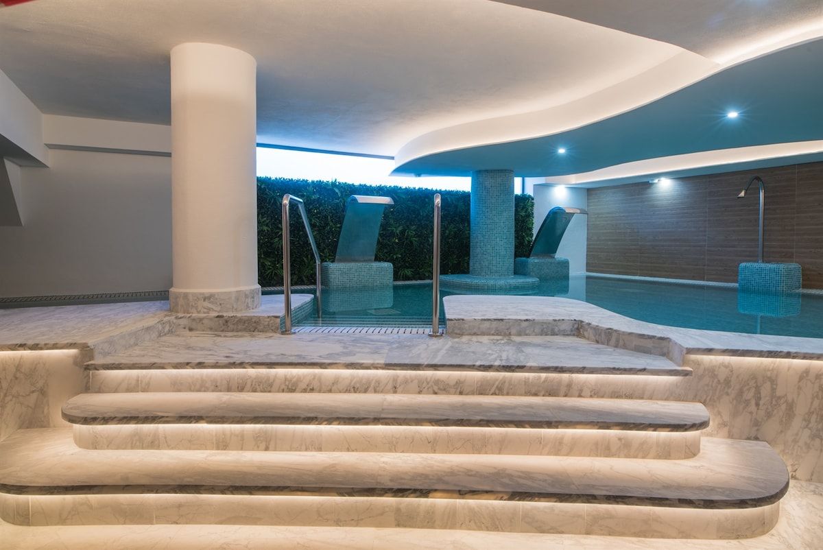 Un espace piscine intérieur moderne, avec des marches menant à un coin salon, le tout entouré d’un jardin aménagé.