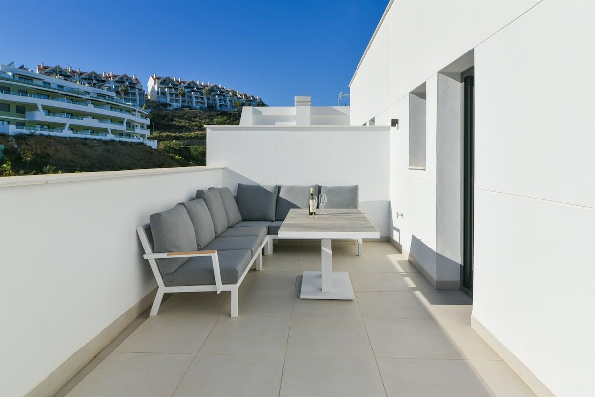 An outdoor patio with a sofa and table, white walls, and a view of a hillside with buildings.