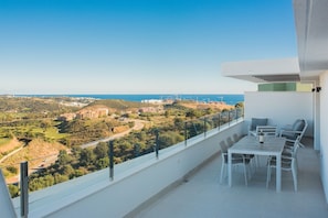 A balcony with a table and chairs, overlooking a coastal landscape with buildings and greenery.