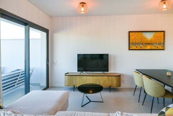 A modern hotel room with a large bed, a TV, a dining table with chairs, and a painting on the wall.