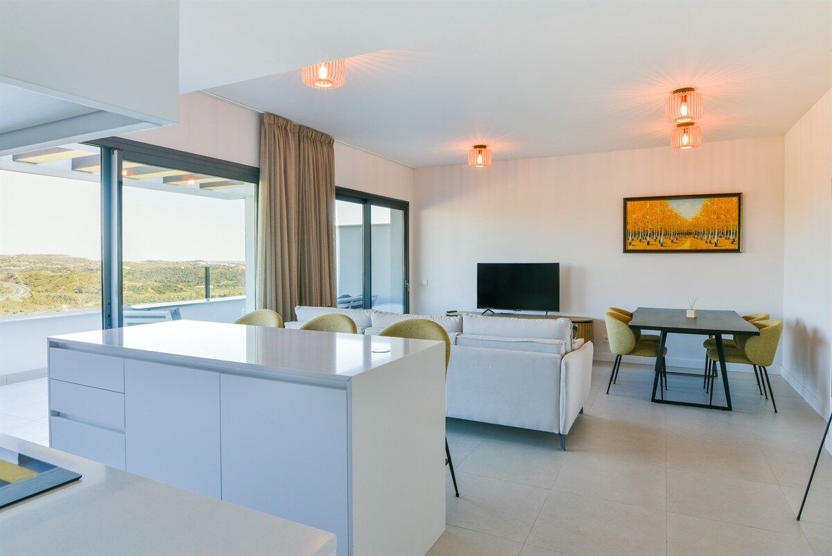 A modern hotel room with a large window, a flat-screen TV, a dining area, and a painting on the wall.