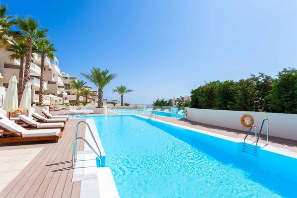 A swimming pool with lounge chairs and umbrellas, surrounded by palm trees and a clear sky.