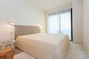 Een slaapkamer met een groot bed, nachtlampjes, een klein tafeltje en een raam met gordijnen.