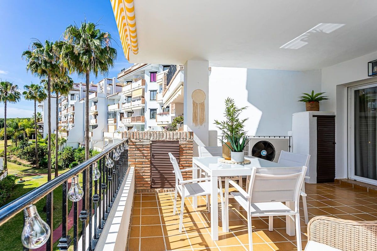 Un balcon avec une table et des chaises, donnant sur un quartier résidentiel bordé de palmiers et de bâtiments.