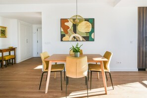 A dining area with a wooden table and four chairs, a hanging light fixture, and a painting on the wall.