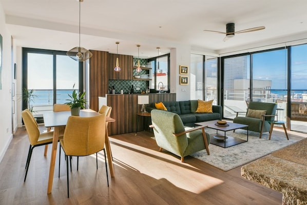 A modern living room with a dining area, a sofa set, and a kitchen area with a view of the sea.