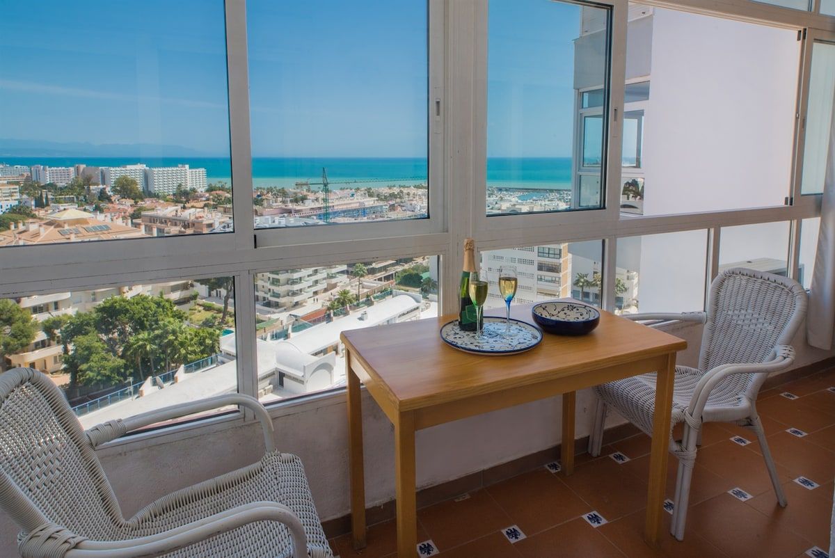 A balcony with a table, two wicker chairs, and a view of the sea and buildings.