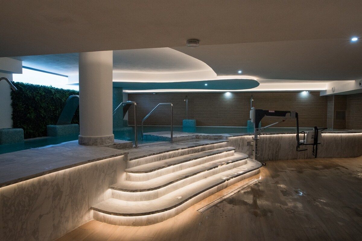 Un espace piscine intérieur moderne, avec des marches menant à l’eau, un coin salon et une ambiance évoquant un spa.