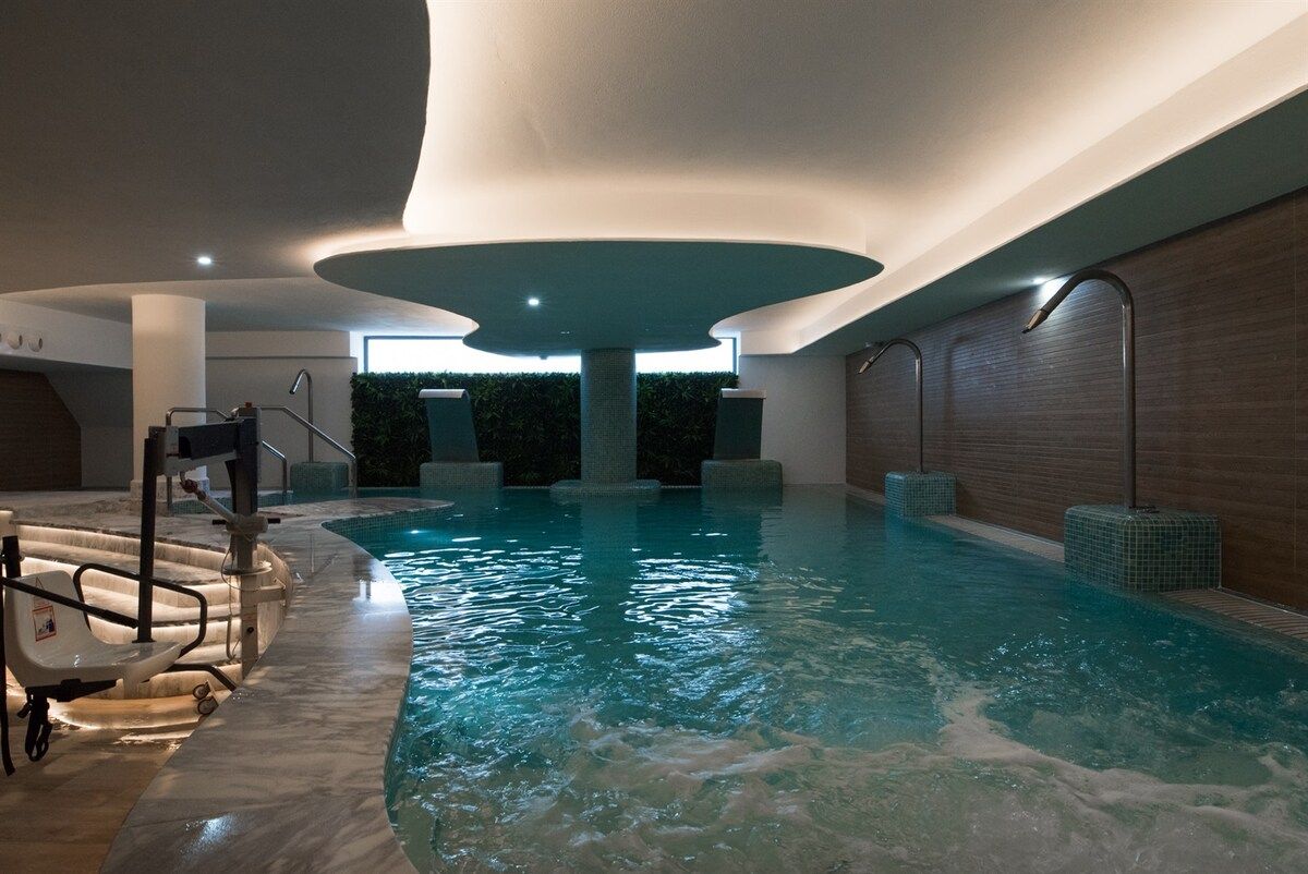 Una piscina interior con una fuente central, rodeada de suelo y paredes con azulejos, y una zona de estar con mesa junto a la piscina.