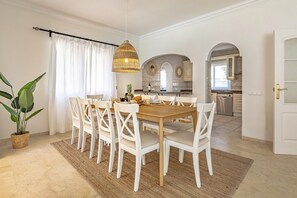 A dining area with a wooden table and white chairs, a hanging light fixture, and a view into the kitchen.