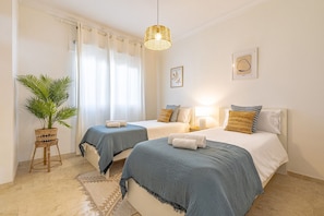 A hotel room with two beds, a potted plant, a woven light fixture, and artwork on the walls.