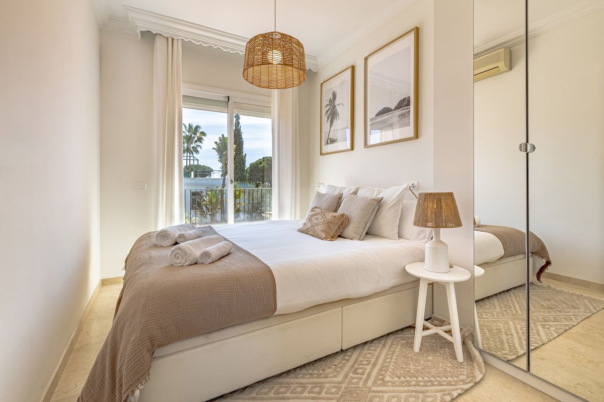 A bedroom with a large bed, a bedside table, a lamp, and a view of the outdoors.