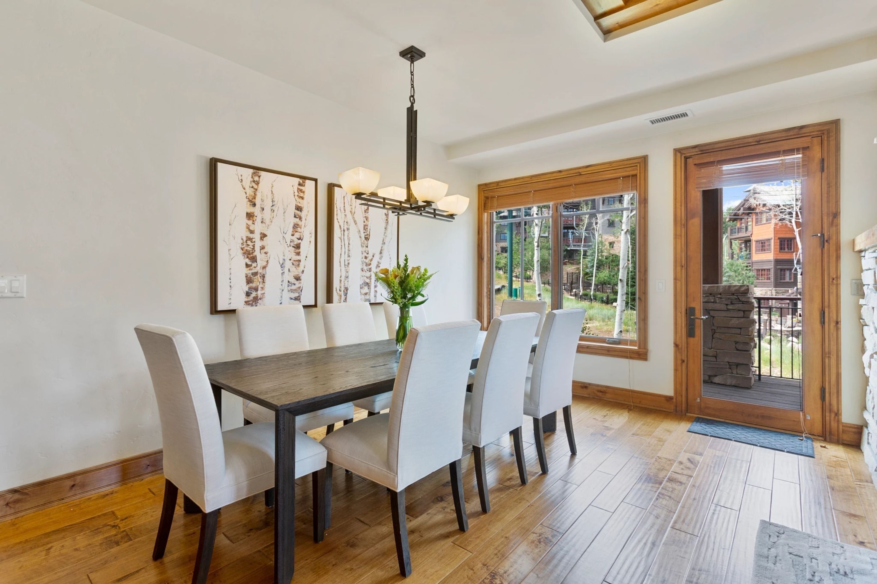 A dining area with a wooden table and six chairs, a chandelier, and a large window with a view of a stone building.