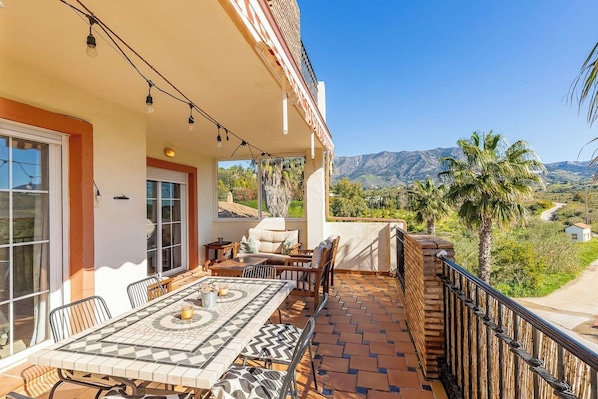 A balcony with a dining table and chairs, a view of mountains, and a palm tree.