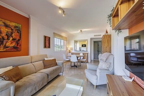 A modern living room with a sofa, armchairs, a glass coffee table, and a TV. There is a dining area with chairs and a table, and a kitchen in the background.