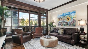 A spacious living room with a large window, leather sofas, a round ottoman, and a painting on the wall.