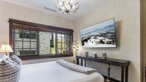 A bedroom with a large bed, a stone wall, a window with blinds, a wooden console table, and a painting of a snowy landscape.