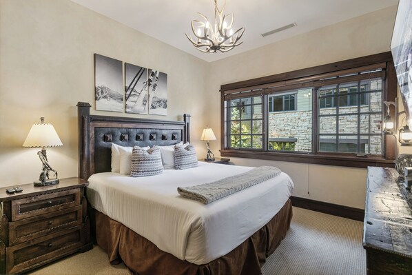 A bedroom with a large bed, two bedside lamps, a chandelier, a stone wall, and a window with a view of a building.