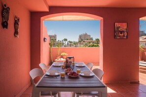 Un comedor con una mesa preparada para dos, vista a edificios y palmeras, y un cactus al fondo.