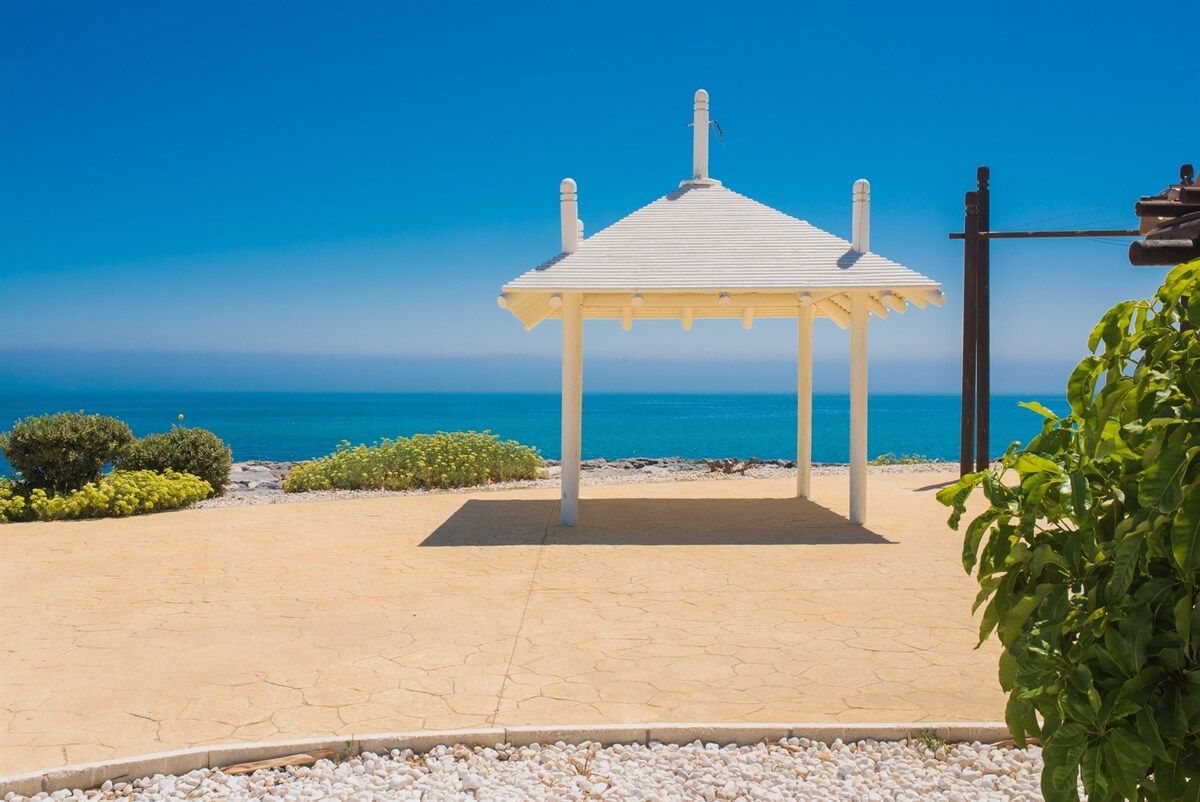 Ein Gazebo mit weißem Dach und Säulen, umgeben von einem Kiesweg und Grünflächen, mit Blick auf das klare, blaue Meer.