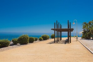 A paved walkway with a wooden pergola structure, benches, and a lamp post, leading to a beach with clear blue water.