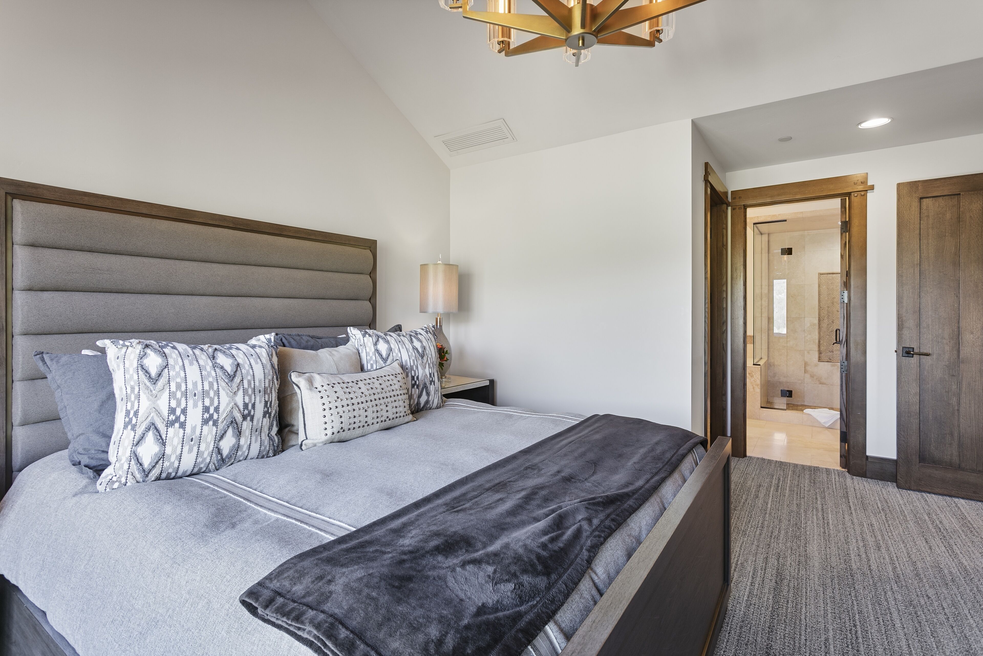 Un dormitorio moderno con una cama grande, cabecera, lámpara de noche y vistas al baño.